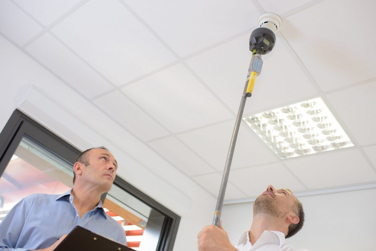 two men inspect a smoke alarm from the ground due to hierarchy of controls